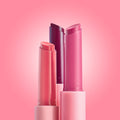 Three She’s So Juicy gloss balm sticks stand uncapped, showing coral, berry, and rosy pink bullets against a soft pink gradient, highlighting the ColourPop So Juicy Plumping Gloss Balm with Peptides for shiny, fuller-looking lips.