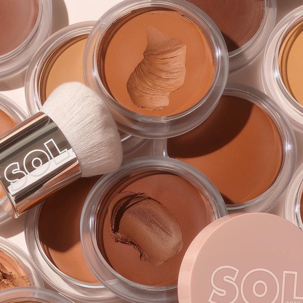 ColourPop SOL Face & Body Bronzing Balm in Medium Dark shows a creamy swipe in an open clear-lid pot, surrounded by other shades and a SOL-branded white kabuki brush in a flat lay on a light background.