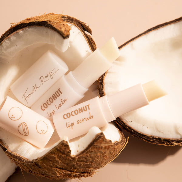 Coco Kisses ColourPop Lip Care Set showing Fourth Ray Beauty coconut lip balm and lip scrub sticks, uncapped and angled, cradled in cracked coconut halves on a soft beige surface, highlighting a tropical, hydrating lip care duo.