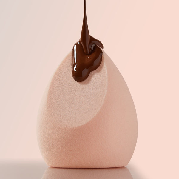Pretty Fresh Complexion Blending Sponge catching a stream of chocolate-brown foundation on its slanted teardrop tip, shown close-up against a soft pink background; a ColourPop Makeup Tools & Accessories essential for seamless base application.