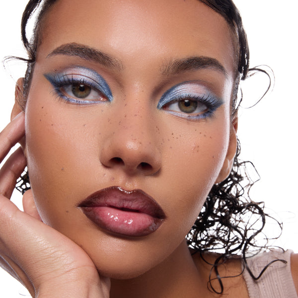 ColourPop Blue Moon shadow palette creating frosty blue shimmer and bold winged eyeshadow on a model’s lids, smoked along the lower lash line, captured in a studio close-up with glossy deep-brown lips and subtle freckles.