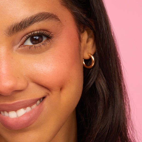 Smiling model’s left cheek and lips wearing Mood 24/7 ColourPop Cheek & Lip Set, showing dewy peach blush and rosy‑nude lip tint with a soft glow; a gold hoop earring and dark hair frame the face against a pink background.