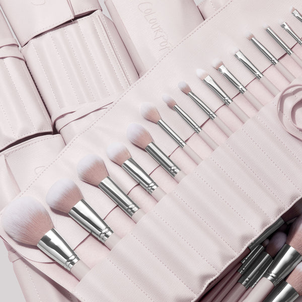 Stone Cold, a ColourPop Makeup Brush Kit, fanned in a blush-pink roll as face and eye brushes slot into stitched pockets, soft white-to-pink bristles and silver ferrules gleaming, with additional matching brush rolls layered behind.