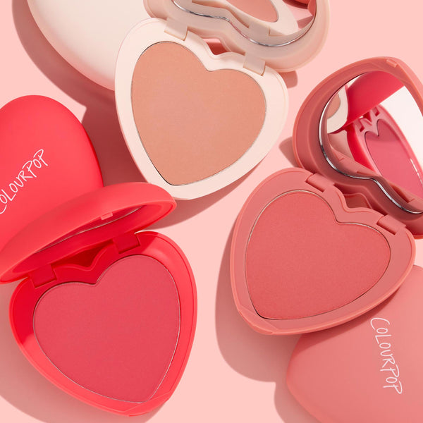 ColourPop Current Crush Heart Powder Blush Set displays open heart compacts with peach, coral, and rosy pink powders and built-in mirrors, arranged on a soft pink backdrop; ColourPop Heart Powder Blush Set for a buildable cheek flush.
