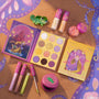 Disney Tangled and ColourPop Collection full makeup set laid flat, featuring Rapunzel-themed eyeshadow palette, lip glosses, glitter, and green chameleon accessory on a purple-and-gold illustrated background promoting ColourPop back in stock collection.