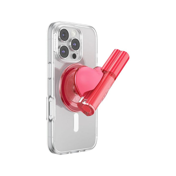 ColourPopSockets ColourPop Lip Stain secured in a pink heart PopSockets grip on a clear smartphone case, the glossy pink tube angled outward against a white background, combining phone grip and lip stain holder for on‑the‑go use.