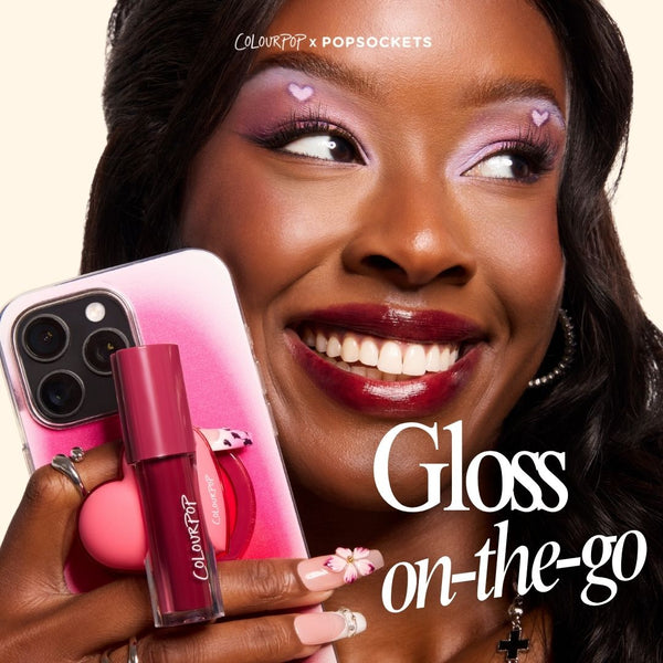 ColourPop Lip Stain tube in deep berry held against a pink smartphone with a ColourPop PopSocket, as a smiling model shows glossy stained lips and lilac heart eye makeup; ColourPopSockets collab highlights on-the-go lip color.