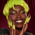 Model smiles with eyes closed, showing shimmery olive green eyeshadow, warm blush, and glossy deep brown lips, styled with a neon Grinch‑green wig against red velvet drapes - highlighting the ColourPop X The Grinch Full Collection Set by ColourPop.