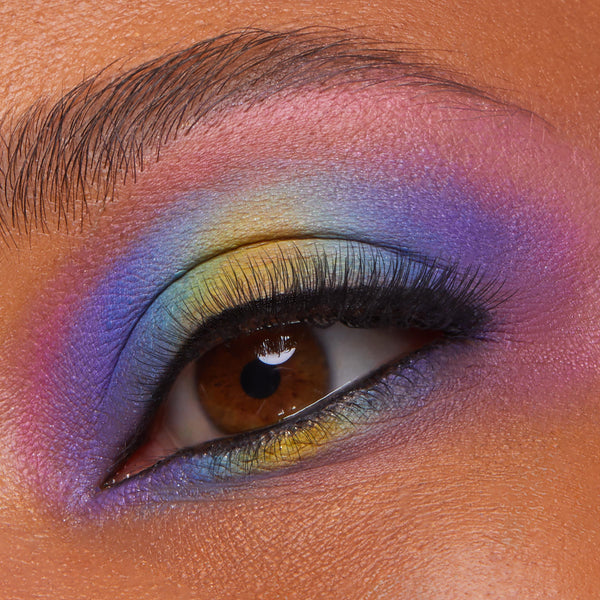 Rainbow gradient eyeshadow arcs across a single eye, blending yellow inner corner into teal, blue, purple, and pink with black liner and full lashes, showcasing the Partner Up ColourPop Shadow Palette from the Shadow Palette & pH Lipstick Set in a studio close-up.