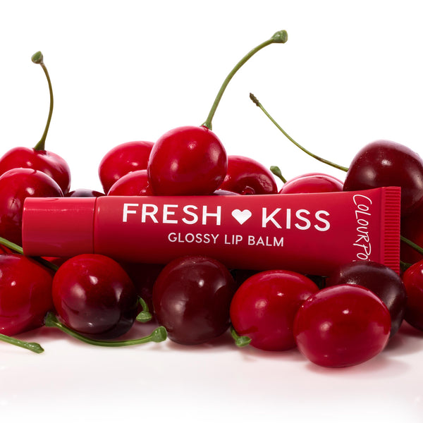 ColourPop Fresh Kiss glossy lip balm tube from the Get Rich ColourPop Glossy Lip Balm Trio resting among ripe cherries, showcasing a cherry-red tint and shiny finish in a studio close-up on a white background.