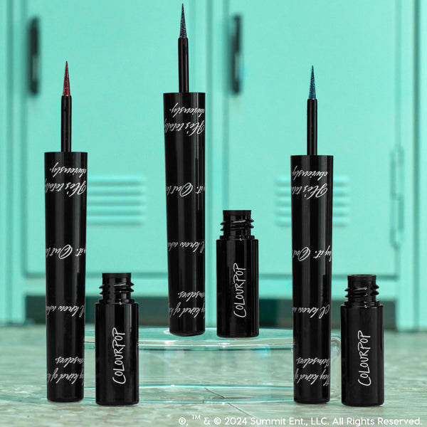 ColourPop Graphix Ink Liner I Know What You Are stands uncapped, showing ultra-fine red, blue, and teal shimmer tips; glossy black tubes with white script sit on a clear riser before mint school lockers for a back-to-school vibe.