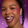 Smiling model wearing Heart to Get Sorry Not Sorry blush and UGL set, showing bold berry cheek color and glossy lips against a vibrant purple background with subtle sparkles.