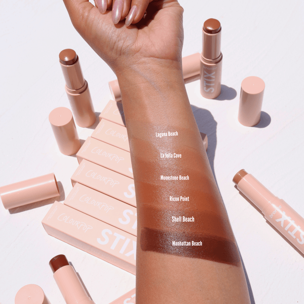 ColourPop Bronzer Stix in Laguna Beach swatched on a forearm - a warm, sun-kissed tan stripe - with additional shade swatches below and nude-pink STIX tubes and boxes scattered on a white surface in bright daylight.