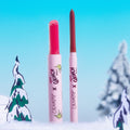 ColourPop Berry Grinchy So Juicy Plumping Gloss Stick stands uncapped with a vivid pink bullet beside a slim neutral lip pencil, both upright in snowy Grinch-themed scenery with fir trees and bright blue sky, collaboration branding on tubes.