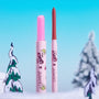 Colourpop Pinky Who So Juicy Plumping Gloss Stick stands uncapped beside a coordinating lip liner, revealing a glittery pink bullet; both in The Grinch x ColourPop pink tubes, set in a snowy pastel-blue holiday scene with cartoon trees.