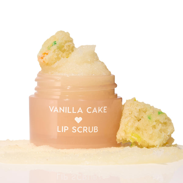 Vanilla Cake ColourPop Lip Scrub jar sits open, sugary scrub mounded on top and swatched at the base, flanked by funfetti cake crumbs on a clean white studio background.