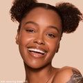 Smiling model wearing ColourPop Pretty Fresh Tinted Foundation Balm shade Dark 20, showcasing an even, dewy finish; close-up face with glossy lips and natural brows against a soft beige studio background.
