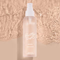 ColourPop Pretty Fresh Setting Spray, Lock It In, stands upright with a white pump on a frosted blush bottle labeled peptides + niacinamide, casting a soft shadow against rippled beige water for a clean, dewy makeup-locking look.