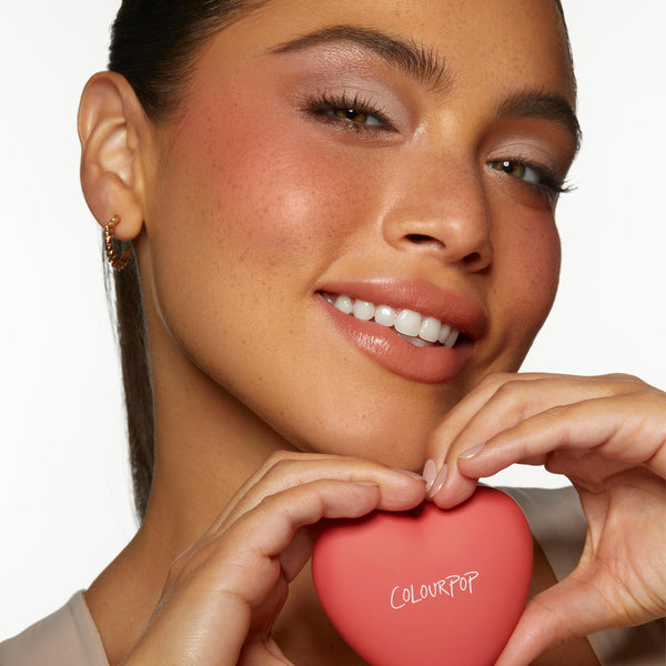 Heart-shaped Cupid Called powder blush compact labeled ColourPop held between a model’s hands forming a heart, showing a soft rosy cheek glow in a studio close-up on white, featuring the ColourPop Heart Powder Blush Set.