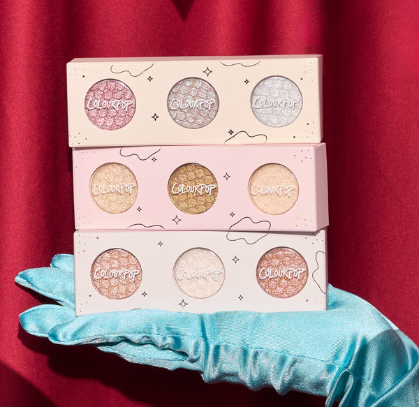 ColourPop Matcha and Mochi Super Shock Shadow trio, three pastel palettes of shimmery eyeshadows stacked on a teal satin glove against a red curtain backdrop, showcasing glittery metallic shades for vibrant eye looks.