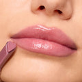 High-shine pink lips wearing Forbidden Love, a ColourPop So Juicy Plumping Gloss, as a heart-tipped mauve applicator glides along the lower lip in a close-up beauty shot, showcasing a cushy, plumped finish and glassy sheen.