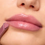 High-shine pink lips wearing Forbidden Love, a ColourPop So Juicy Plumping Gloss, as a heart-tipped mauve applicator glides along the lower lip in a close-up beauty shot, showcasing a cushy, plumped finish and glassy sheen.