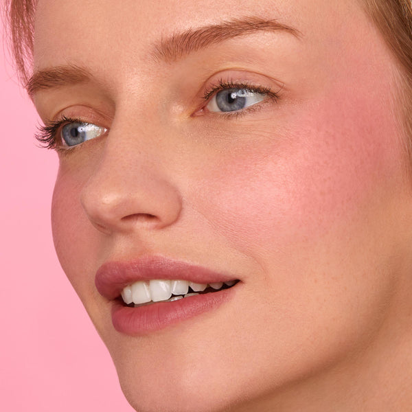Rosy cheek and soft pink lips glow, showcasing the That’s So Rosy ColourPop Cheek & Lip Set on a close-up model; creamy blush and matching lip tint blend seamlessly with a dewy finish against a pastel pink background.
