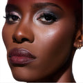 Model wearing Change Me Mega Highlighter from the Twilight collection, showing a radiant glow on deep skin with shimmering cheekbone highlight, metallic smoky eye makeup, and berry lipstick against a clean white background.