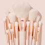 Ultimate Brush Roll, a ColourPop Makeup Brush Kit, fanned upright with soft white bristles, rose-gold ferrules, and cream handles, showcasing face and eye brushes for powder, blush, and eyeshadow blending on a blush-pink studio backdrop.