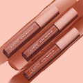 Three ColourPop Ultra Matte Lip tubes in the Warmed Up bundle rest diagonally atop wide nude‑brown swatches, showcasing a full‑coverage ultra matte finish with clear barrels and blush pink star caps on a peach backdrop.