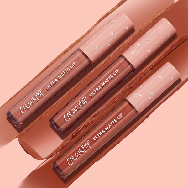 Three ColourPop Ultra Matte Lip tubes in the Warmed Up bundle rest diagonally atop wide nude‑brown swatches, showcasing a full‑coverage ultra matte finish with clear barrels and blush pink star caps on a peach backdrop.