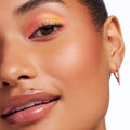 Vibe Shift ColourPop Chrome Liquid Shadow sweeping across the eyelid in a pink-to-gold metallic shift, with defined lashes, glossy nude lips, and a gold geometric hoop earring in a clean white studio close-up.