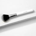 ColourPop Large Powder Brush with fluffy black-to-white bristles rests diagonally on a white textured block, showcasing a chrome ferrule and white handle logo, a soft full-size makeup brush for sweeping on setting or finishing powder.