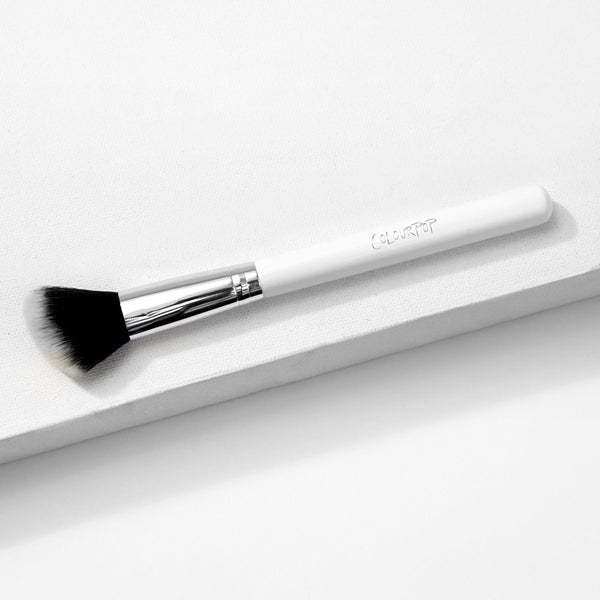 ColourPop Angled Face Brush makeup brush resting diagonally on a white textured platform, featuring a white handle with ColourPop logo, shiny silver ferrule, and soft black-to-white angled bristles for precise blush, bronzer, and contour.