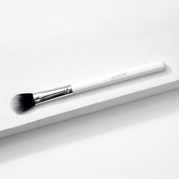 ColourPop Small Fluff Brush lies diagonally on a white textured block, showing soft synthetic black-to-white bristles, a chrome ferrule, and a white handle; a versatile makeup brush for precise blending in a clean studio setting.