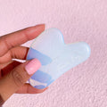 Opalite Crystal Gua Sha facial massage tool held in a hand, showing its iridescent blue‑white stone and curved edges, branded Fourth Ray Beauty, against a pink textured background; part of ColourPop Skincare Tools And Accessories.