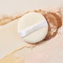 Powder Puff pressing into loose setting powder, a soft round ivory pad with COLOURPOP logo strap resting on swirls of beige, tan, and pink powders - an essential from ColourPop Makeup Tools & Accessories for smooth blending and shine control.