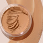 ColourPop SOL Face & Body Bronzing Balm in shade Light sits in a clear round jar with creamy swirls, with warm golden-tan balm swatched around it on a beige surface, showcasing a smooth, blendable texture and natural dewy finish.