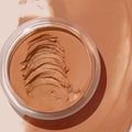 ColourPop SOL Face & Body Bronzing Balm in Medium Dark, creamy bronzer in an open clear jar, shows layered spatula swipes revealing smooth texture, set against broad bronze swatches on a neutral background.