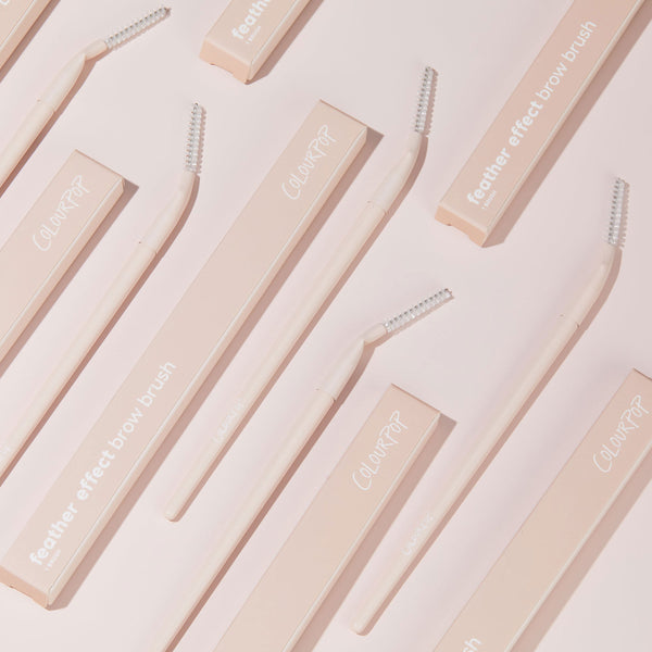 ColourPop Brow Brush with soft pink handle and clear spoolie head, arranged diagonally on a pale pink backdrop beside matching feather effect brow brush boxes, showcasing a makeup brush for shaping and feathering eyebrows.