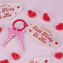 Flirty Talk keychain with two pink leopard-print keys on a red ring rests beside a pearly motel-style tag reading “Talk Flirty To Me,” surrounded by red sugar-coated heart candies on a pastel pink backdrop; ColourPop Tools and Accessories.