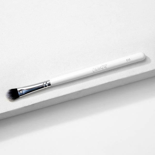 Large Shader Brush makeup brush by ColourPop lies diagonally on a white textured surface, showcasing a white handle with logo and E13, a shiny silver ferrule, and flat dense black-to-white bristles designed for packing and shading eyeshadow.