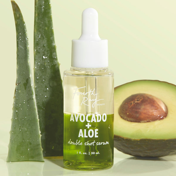 Dropper bottle of Fourth Ray Beauty Avocado + Aloe Double Shot face serum (1 fl oz) stands upright, showcasing its bright green formula, flanked by dewy aloe blades and a halved avocado on a pale green backdrop; a nourishing ColourPop Face Serum.