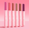 Six ColourPop So Juicy Plumping Gloss Balm with Peptides sticks stand upright, caps off to reveal pink, coral, nude, caramel, terracotta, and deep berry shades in matte pink tubes labeled SO JUICY, lined on a pink gradient background - So Balm bundle.
