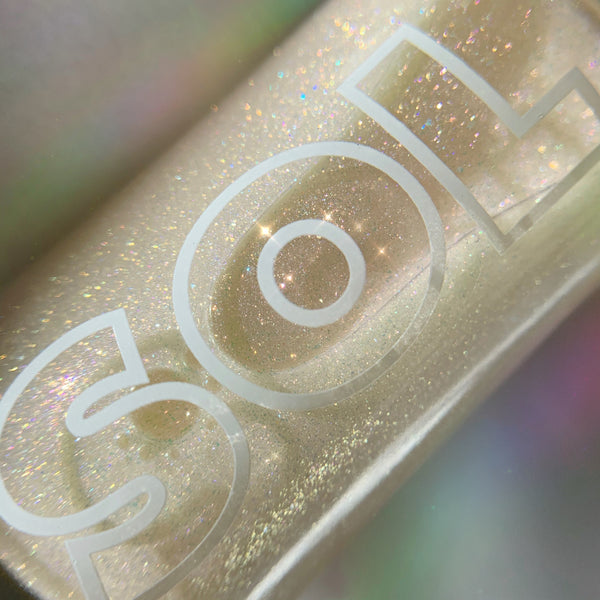 SOL-labeled bottle sparkles as fine rainbow glitter swirls in a golden base; ColourPop SOL Shimmering Dry Oil, Rainbow Glow, shown in a tight macro against a soft iridescent backdrop for a luminous body sheen.