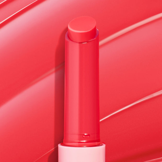 Delulu shade of the ColourPop So Juicy Plumping Gloss Balm with Peptides, twist-up bullet extended upright from a sleek tube with a light pink base, glossy coral-red stick centered against a swirled pink backdrop.