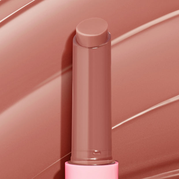 ColourPop So Juicy Plumping Gloss Balm with Peptides in Doe stands uncapped, twist-up stick revealing a creamy warm nude-brown tint, set against matching glossy swipes, in a pink tube for a plumping, glossy finish.