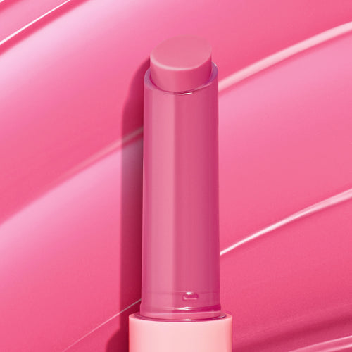 Dolled Up pink twist-up lip balm stick showcasing the ColourPop So Juicy Plumping Gloss Balm with Peptides, standing uncapped to reveal a creamy rosy tint against glossy pink swatch strokes that emphasize shine and a plumping finish.