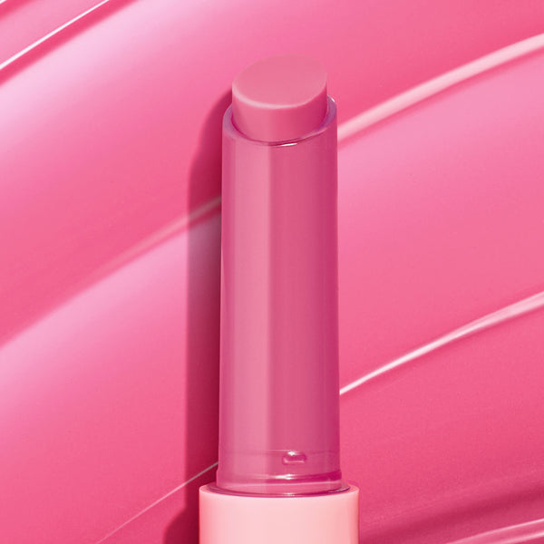 Dolled Up pink twist-up lip balm stick showcasing the ColourPop So Juicy Plumping Gloss Balm with Peptides, standing uncapped to reveal a creamy rosy tint against glossy pink swatch strokes that emphasize shine and a plumping finish.