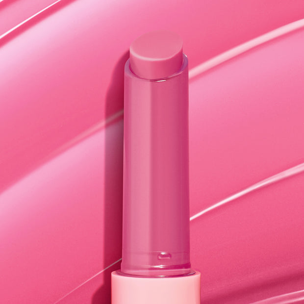 Dolled Up pink twist-up lip balm stick showcasing the ColourPop So Juicy Plumping Gloss Balm with Peptides, standing uncapped to reveal a creamy rosy tint against glossy pink swatch strokes that emphasize shine and a plumping finish.
